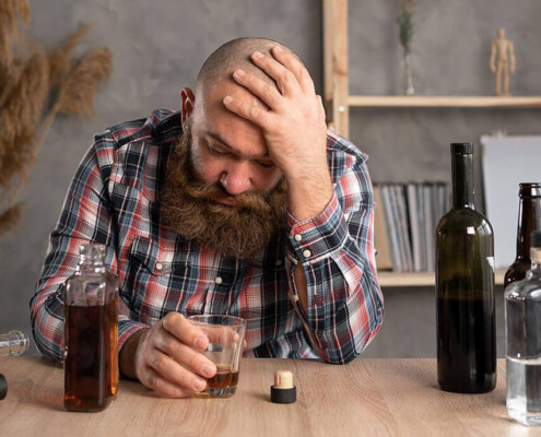 A man holds his head while sitting at a table with multiple liquor bottles. A psychiatrist in New York, NY can offer alcohol addiction help in New York, NY. Learn more about how alcohol addiction treatment in New York, NY can support you today!