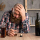 A man holds his head while sitting at a table with multiple liquor bottles. A psychiatrist in New York, NY can offer alcohol addiction help in New York, NY. Learn more about how alcohol addiction treatment in New York, NY can support you today!