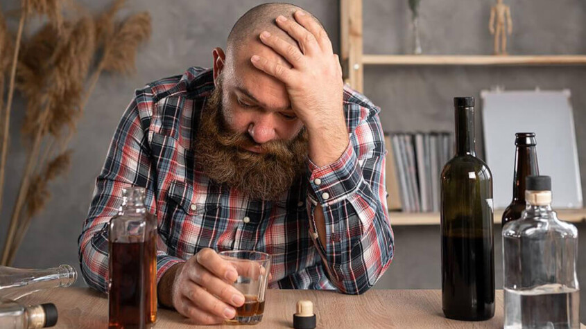 A man holds his head while sitting at a table with multiple liquor bottles. A psychiatrist in New York, NY can offer alcohol addiction help in New York, NY. Learn more about how alcohol addiction treatment in New York, NY can support you today!