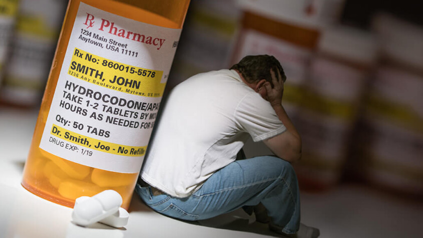 opioid addiction - man sitting beneath the shadow of a large bottle of prescription opioids