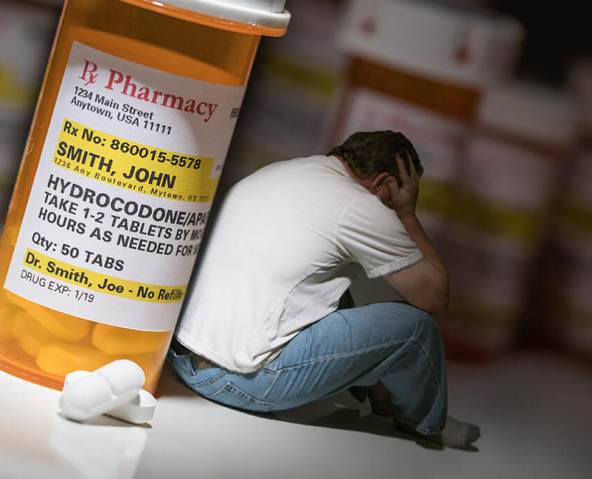 opioid addiction - man sitting beneath the shadow of a large bottle of prescription opioids