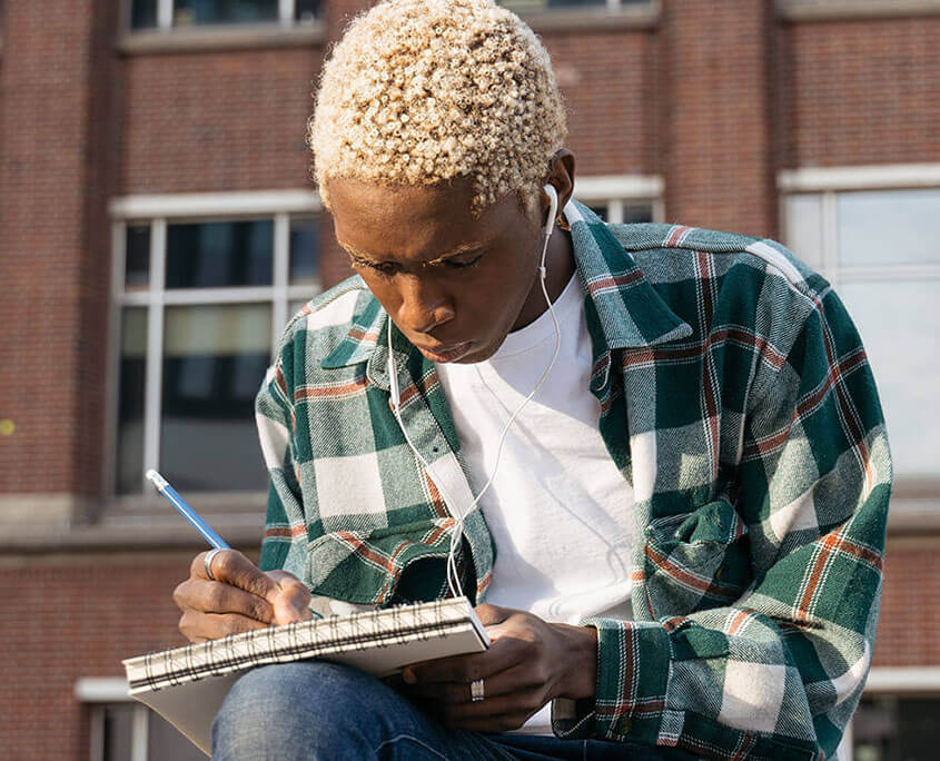 college student outside listening to lecture on mobile device and taking notes. A teenage psychiatrist in New York, NY can offer support with teenage psychiatry and other services. Contact a psychiatrist for young adults to learn about young adult psychiatry in New York, NY today!