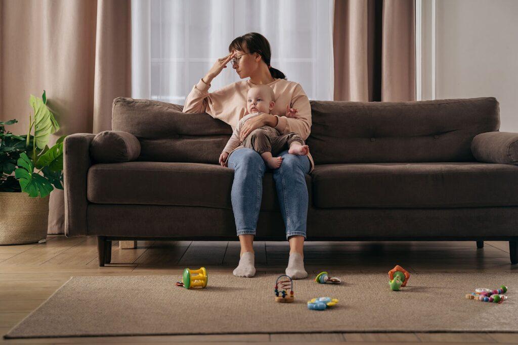 An image of a woman holding her baby sitting on a couch surrounded by toys. The stress of motherhood is not one that can easily be coped with, before slipping into addiction abuse, reach out to us for Alcohol addiction help in New York, NY!