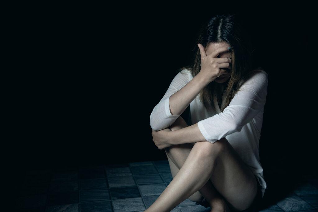A woman covers her face while sitting in a dark room. This could represent the isolation of trauma a PTSD therapist in New York, NY can offer support in overcoming. Learn more about PTSD treatment in NYC, Manhattan by searching “trauma therapist NYC, Manhattan” today. 

