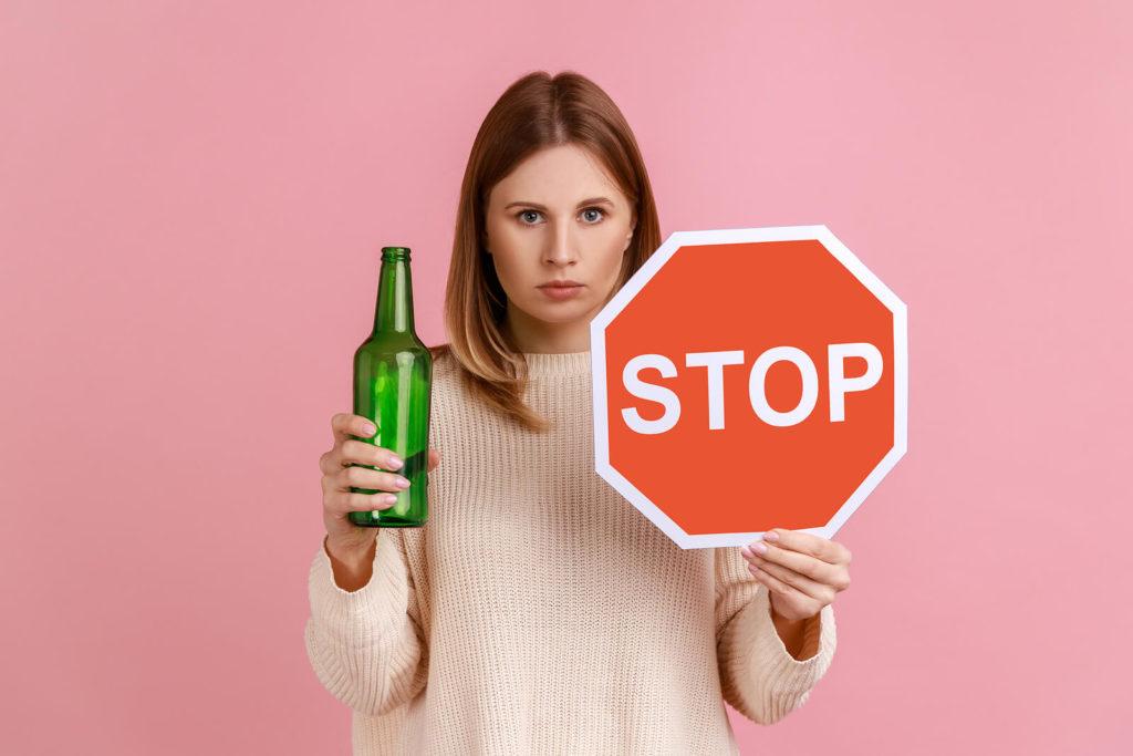 A woman holds a bottle in one hand and a stop sign in the other. Learn how alcohol addiction treatment in New York, NY can offer support today with a trained psychiatrist. Learn more about alcohol treatment in New York, NY by searching "telepsychiatry in New York, NY today."