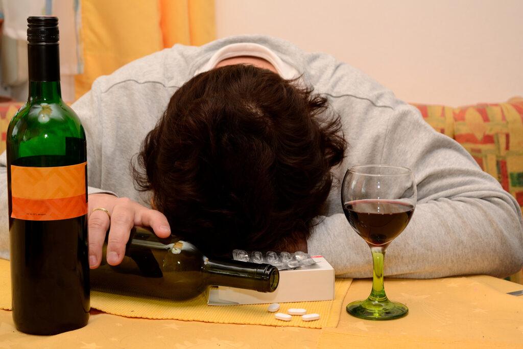 A man lays with his head on a table next to wine bottles and pills. This could represent the struggles of relapse. Learn how an addiction psychiatrist in New York, NY can offer alcohol addiction help in New York, NY, and other services. 
